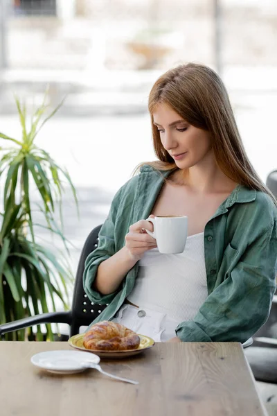Pelirroja sosteniendo taza de café cerca de sabroso croissant en la terraza de la cafetería - foto de stock