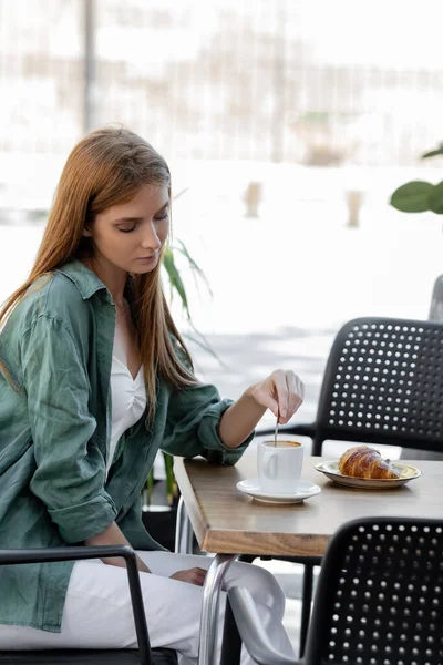 Рыжая женщина, мешающая кофе ложкой возле вкусного круассана на террасе кафе — стоковое фото
