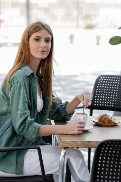 Mujer con cabello rojo revolviendo café con cuchara cerca de sabroso croissant en la terraza de la cafetería - foto de stock