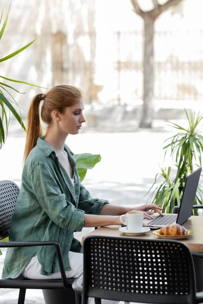 Вид сбоку женщины с рыжими волосами на ноутбуке возле чашки кофе и вкусного круассана на террасе кафе — стоковое фото
