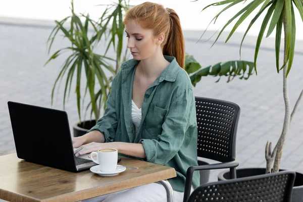 Young woman with red hair using laptop near cup of coffee in cafe terrace — Stock Photo