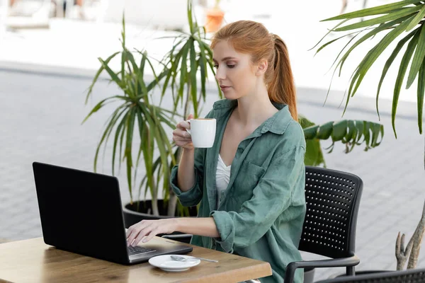 Young woman with red hair using laptop and holding cup of coffee on summer terrace — Stock Photo