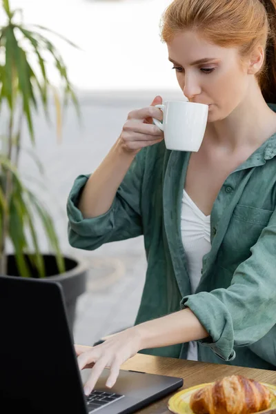 Young woman with red hair using laptop and drinking coffee in cafe terrace — Stock Photo