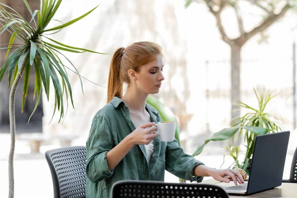 Pigiste rousse à l'aide d'un ordinateur portable et tenant une tasse de café sur la terrasse d'été — Photo de stock