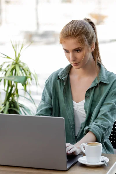 Focused woman with red hair using laptop near cup of coffee in cafe terrace — Stock Photo