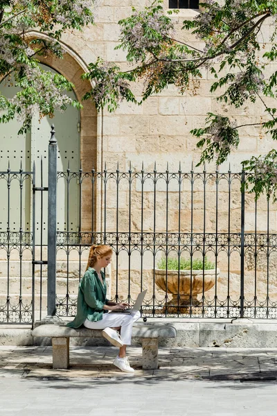 Рыжая женщина сидит на бетонной скамейке и использует ноутбук возле кованого забора в Валенсии — стоковое фото