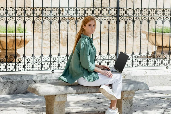 Rossa freelance seduta su panchina in cemento con laptop vicino recinto forgiato in valencia — Foto stock