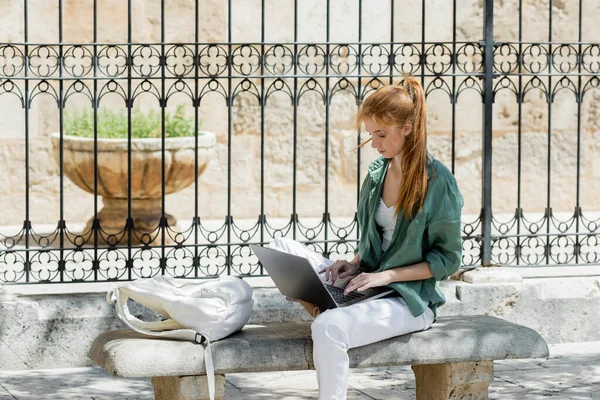 Рыжая молодая женщина сидит на бетонной скамейке и использует ноутбук возле кованого забора — стоковое фото