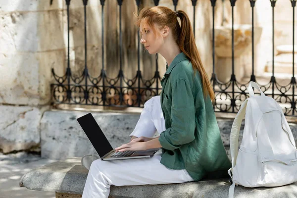 Pelirroja freelancer sentada en banco de hormigón y usando laptop con pantalla en blanco cerca de valla forjada en valencia - foto de stock