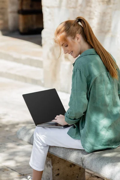 Un pigiste souriant assis sur un banc en béton et utilisant un ordinateur portable avec écran vierge — Photo de stock