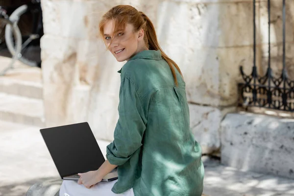 Cheerful freelancer sitting on concrete bench and using laptop with blank screen — Stock Photo