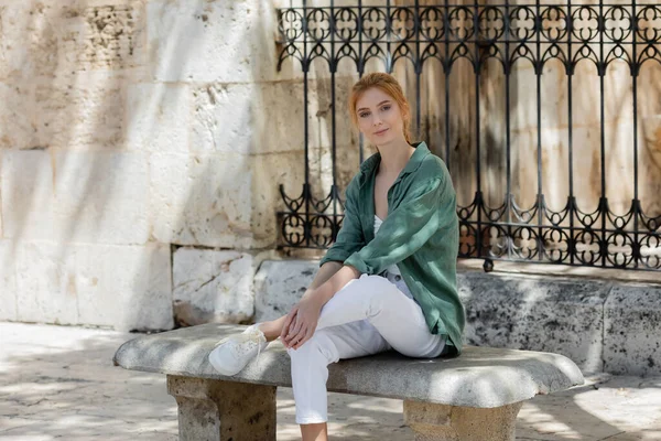 Young woman with red hair in green linen shirt sitting on concrete bench near forged fence — Stock Photo