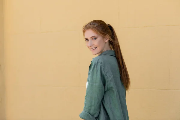 Redhead and happy woman in green linen shirt smiling near beige wall — Stock Photo