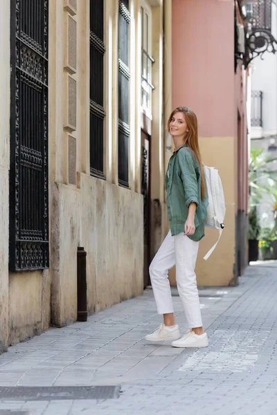 Joven pelirroja viajero con mochila sonriendo mientras camina por la calle de valencia - foto de stock