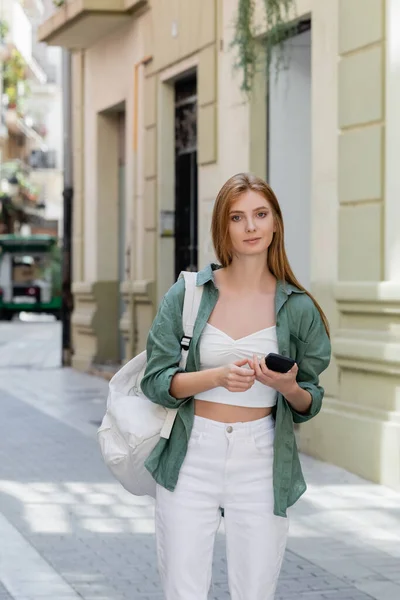 Young redhead traveler with backpack holding smartphone on urban street of valencia — Stock Photo