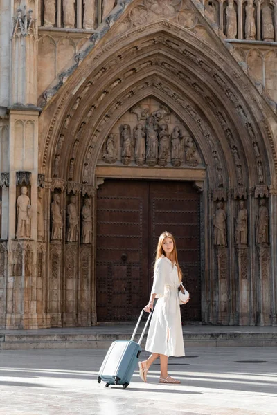 Повна довжина молодої жінки в сукні тримає гаманець і стоїть з багажем у Валенсії — стокове фото