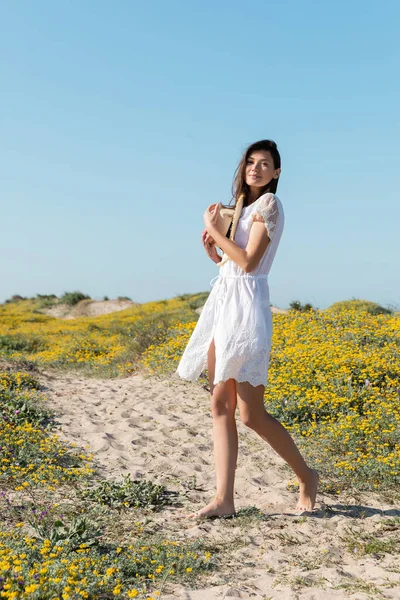 Descalza mujer en vestido sosteniendo sombrero de paja y mirando a la cámara en la playa - foto de stock