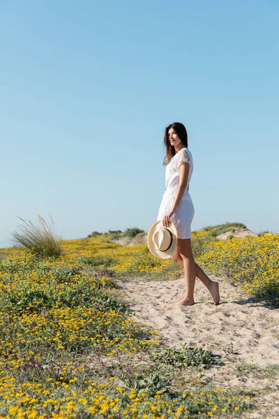 Mujer morena sosteniendo sombrero de paja y mirando hacia otro lado cerca de flores en la playa - foto de stock