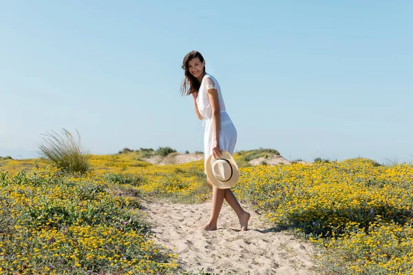 Femme positive en robe tenant chapeau de paille et regardant la caméra sur la plage — Photo de stock