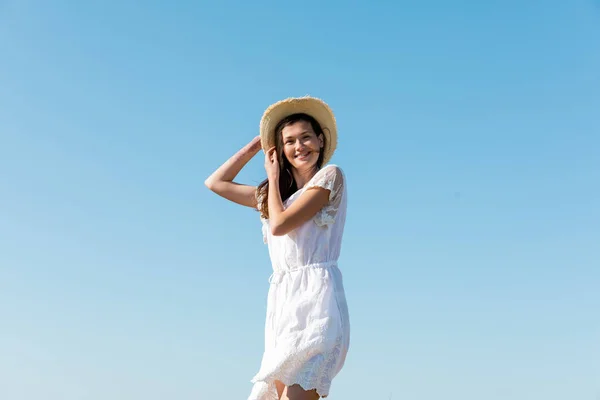 Vue à angle bas de la femme souriante en chapeau de soleil et robe regardant la caméra avec le ciel à l'arrière-plan — Photo de stock