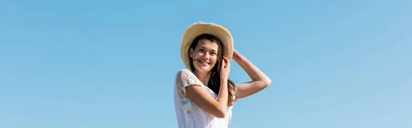 Vue à angle bas de jolie femme en chapeau de soleil souriant à la caméra avec ciel bleu à l'arrière-plan, bannière — Photo de stock