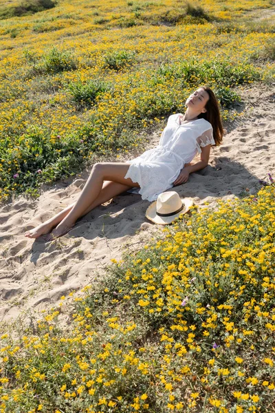 Красивая брюнетка лежит рядом с соломенной шляпой на песчаном пляже рядом с цветами — стоковое фото