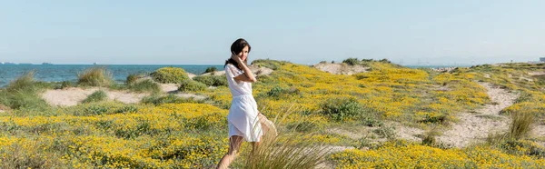 Hübsche Frau im Sommerkleid blickt in die Kamera in der Nähe von Blumen am Strand, Banner — Stockfoto