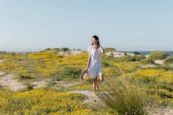 Donna felice con cappello da sole che cammina vicino ai fiori sulla spiaggia — Foto stock