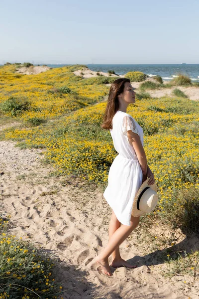 Vista lateral de mujer en vestido sosteniendo sombrero de paja y mirando al mar en la playa - foto de stock