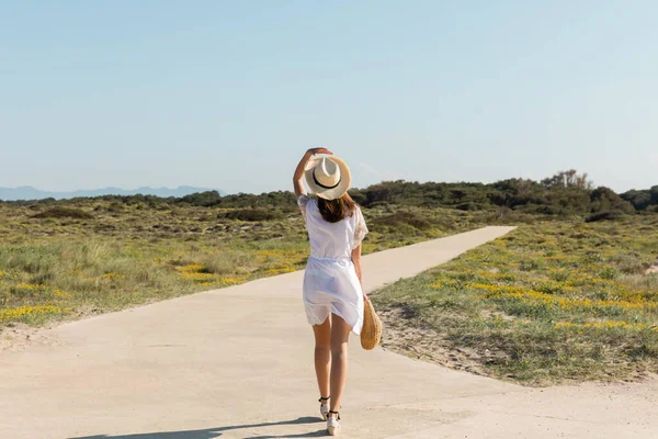 Rückansicht einer Frau mit Sonnenhut und Kleid, die Handtasche hält, während sie am Strand spaziert — Stockfoto