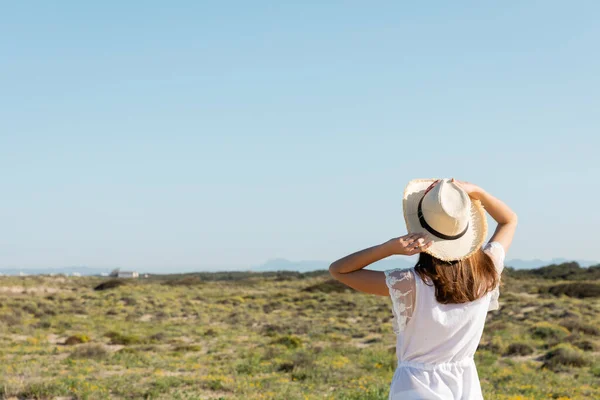 Vista posteriore della giovane donna che tiene il cappello di paglia vicino all'erba sulla spiaggia sullo sfondo — Foto stock