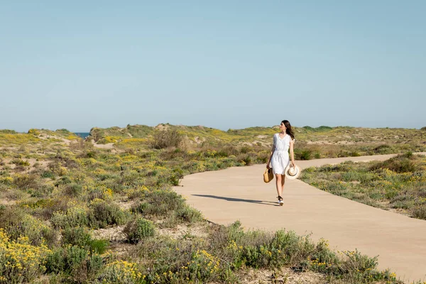 Вид сбоку на симпатичную женщину с сумкой и соломенной шляпой, идущую по тропинке на пляже — стоковое фото