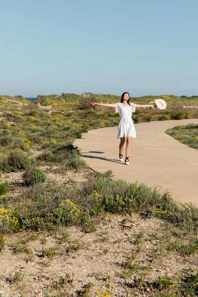 Frau im Kleid schließt Augen und hält Strohhut am Strand — Stockfoto