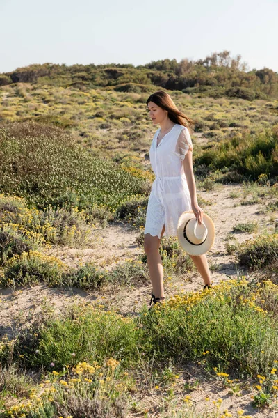 Pretty young woman holding sun hat while walking near plants on beach — Stock Photo