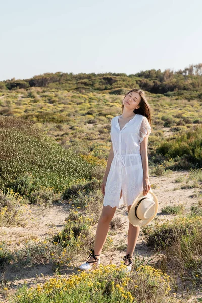 Jeune femme en robe fermant les yeux et tenant un chapeau de paille tout en se tenant sur la plage — Photo de stock