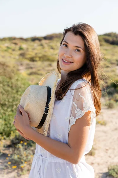 Portrait of young woman holding sun hat and smiling at camera on beach — Stock Photo