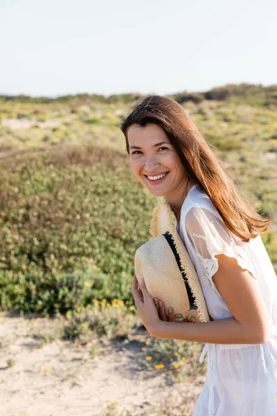 Positive woman in summer dress holding straw hat on blurred beach — Stock Photo