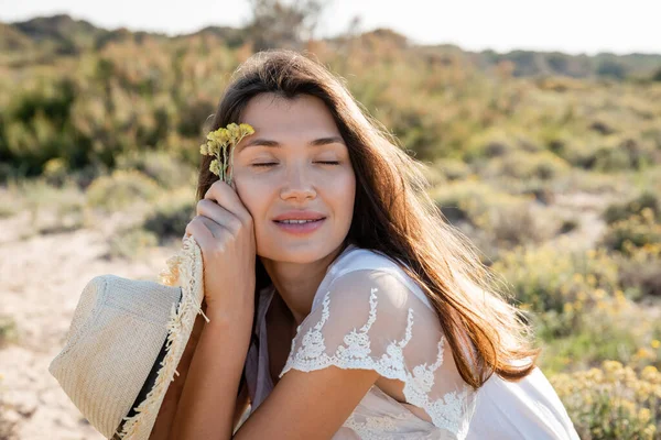 Улыбающаяся женщина с закрытыми глазами держит шляпу солнца и цветы на газоне — стоковое фото