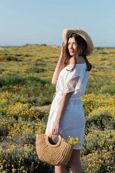 Donna in abito e cappello da sole in possesso di borsetta con fiori e guardando la fotocamera sulla spiaggia — Foto stock