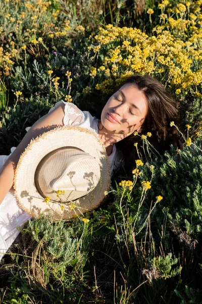 Mujer morena tendida cerca de sombrero de sol y flores en el prado - foto de stock