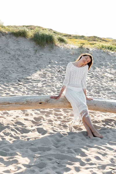 Hübsche Frau im weißen Strickkleid sitzt auf Holzklotz am Strand — Stockfoto