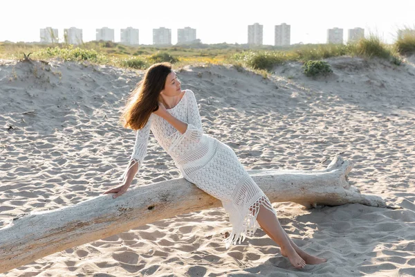 Mujer de pelo largo en vestido sentado en tronco de madera en la playa - foto de stock