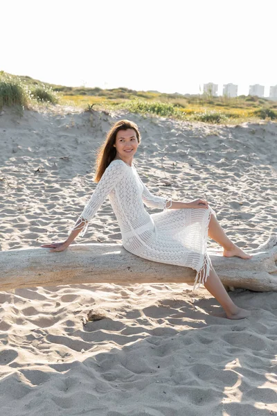 Donna positiva in abito estivo guardando lontano mentre seduto su tronco di legno sulla spiaggia — Foto stock