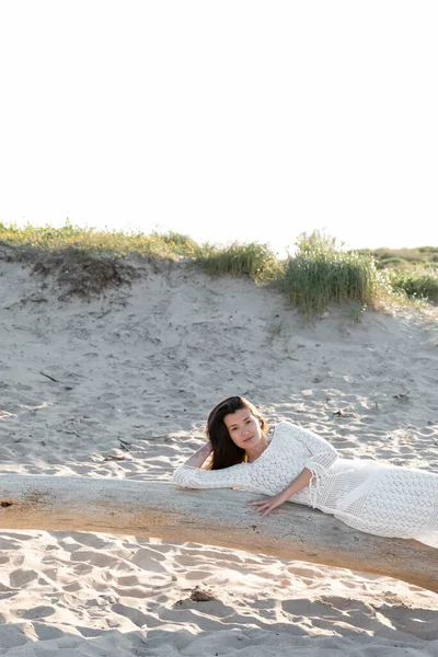 Femme en robe tricotée regardant la caméra alors qu'elle était allongée sur une bûche en bois sur la plage — Photo de stock