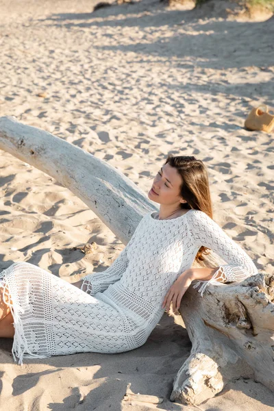 Hübsche junge Frau im Strickkleid sitzt auf Sand in der Nähe von Holzbalken am Strand — Stockfoto