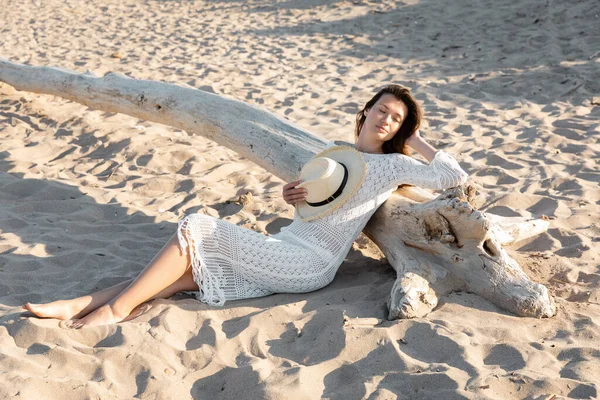 Mujer joven en vestido sosteniendo sombrero de sol mientras descansa cerca de tronco de madera en la playa de arena - foto de stock