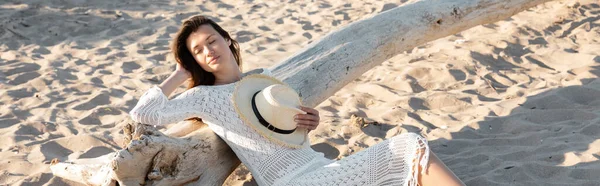 Young woman holding straw hat while relaxing near wooden log on beach, banner — Stock Photo