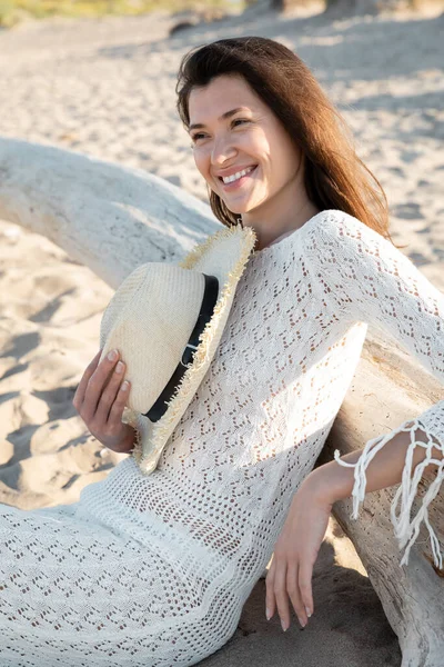 Ritratto di giovane donna in abito lavorato a maglia che tiene il cappello vicino al tronco di legno sulla spiaggia — Foto stock