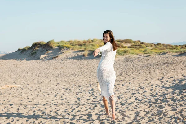 Mulher positiva em vestido de malha olhando para a câmera enquanto caminhava na praia — Fotografia de Stock