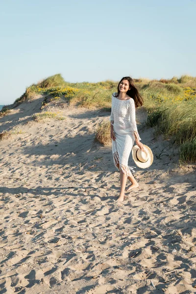 Femme brune positive en robe tenant un chapeau de paille tout en marchant sur une plage de sable — Photo de stock